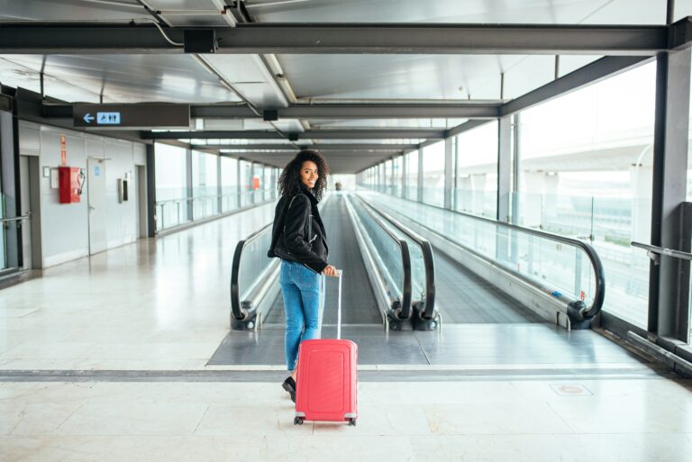 Mulher sorridente caminhando em um terminal de viagens com sua mala de rodinhas e transportando uma impressora portátil compacta.