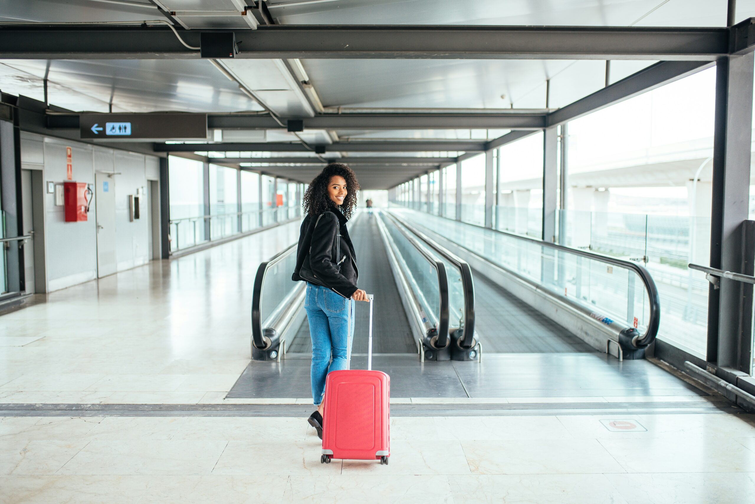 Mulher sorridente caminhando em um terminal de viagens com sua mala de rodinhas e transportando uma impressora portátil compacta.
